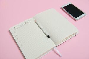 Still life. A white smartphone lying down on pink background next to a pencil on the middle of an open agenda, diary, notebook with a list to do on white sheet of paper in line with copy space photo