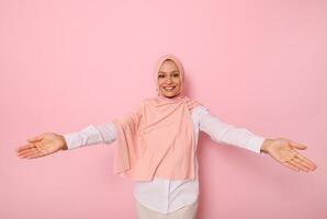 Friendly, cute smiling with a beautiful charming toothy smile young Muslim Arab woman in pink hijab looking at camera with outstretched arms showing hospitality, on colored background with copy space photo