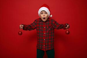 Adorable amazed preteen boy, surprised beautiful child stands on a red background with Christmas spherical shiny toys in his arms, looks at the camera with surprise. New year concept with copy space photo