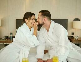 Cheerful young loving married couple, newlyweds sitting on the bed after waking up and enjoying delicious breakfast for two in a bedroom of a luxury hotel while celebrating their marriage anniversary photo