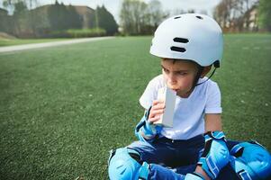 Cute boy in a skateboard helmet and protective equipment resting sitting on a green grass and drinking juice photo