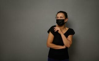Isolated portrait on gray background of a pensive smiling cheerful woman in black medical mask with crossed arms looking thoughtfully to the side at copy space for advertisement. Black Friday concept photo