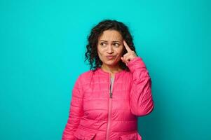 Pensive puzzled thoughtful curly haired woman in bright pink jacket holding her finger on her temple and looking thoughtfully aside to a copy space for advertising text, isolated on blue background photo