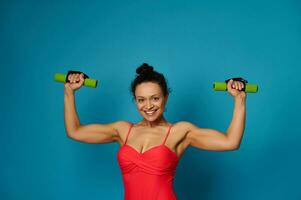 Smiling young athletic woman posing with dumbbells on blue background with copy space photo