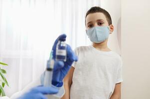 Boy in medical mask looking at the hands of doctor holding a vial with vaccine and syringe on blurred foreground photo