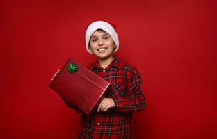 Adorable boy in Santa hat and plaid shirt holds Christmas present in glitter wrapping gift paper with green bow, smiles with toothy smile looking at camera, poses over colored background. Copy space photo