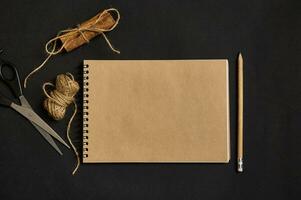 Top view of a craft notepad with an empty blank paper sheet with copy space, a wooden pencil, tied cinnamons with a rope and scissors lying down on black background photo