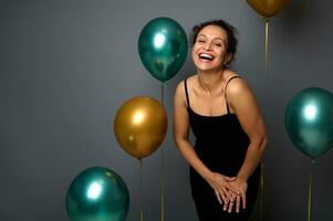 Portrait on gray background with copy space of beautiful woman in evening black dress posing against gold and green air balloons, smiles toothy smile . Birthday and Christmas concept for advertising photo