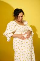 Gorgeous pregnant woman in sundress putting hands on her big belly, making a shape of heart, isolated yellow background photo