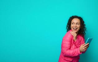 Joyful awesome attractive pretty woman with curly hairstyle, wearing bright pink jacket smiles toothy smile holding smartphone in hand and looks through her shoulders on copy space of blue background photo