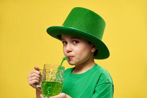 pequeño chico mira a el cámara mientras Bebiendo verde bebida desde un Paja con trébol hoja ornamento celebrando un Santo patrick's día. Copiar espacio foto