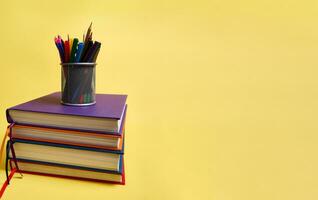 un metal lápiz poseedor con colegio suministros en pilas de multicolor libros. del maestro día concepto, literario, conocimiento, espalda a escuela, educación. amarillo antecedentes Copiar espacio foto