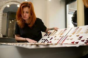 Reflection in the mirror of a concentrated stylish beautiful redhead woman with tattooed arms, professional hairdresser with a palette of different colors to hair dye at hairdressing salon. photo