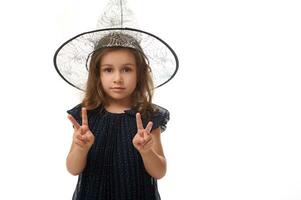 Isolated waist length portrait of pretty little witch girl wearing a wizard hat and dressed in stylish carnival dress, gesturing, showing a peace sign with fingers. Halloween concept photo