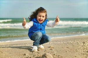 Happy and joyful little girl showing thumbs up while squatting on the beach. International Children's Day. Happy childhood concept. Love nature and planet earth protection concept photo