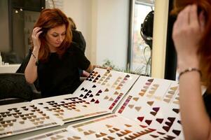 Beautiful fashionable redhead woman, mid adult professional female hairdresser examines palette of hair dye samples while sitting in front of her mirror reflection in a beauty salon photo