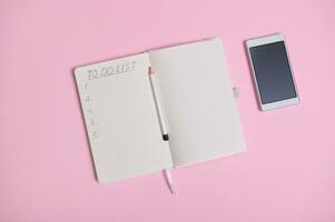 Flat lay of a white mobile phone lying down on pink background next to a pencil on the middle of an open agenda, diary, notebook with a list to do on white sheet of paper in line with copy space photo