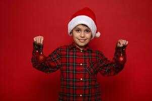 Adorable preadolescent boy, beautiful child poses against colored red background with Christmas spherical shiny toys on his hands, cutely smiles looking at camera. New Year concept with copy space photo