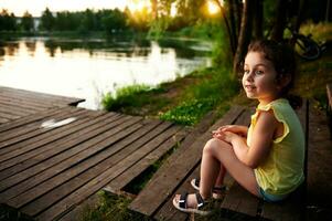 un encantador niña descansando en el muelle y mirando dentro el distancia en contra el fondo de el río banco. cámping, verano temas foto