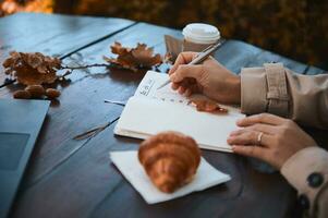 Close-up of hand of a cropped woman holding silver pen and writing the list to do in notepad, while sitting at a wooden table with fallen dry oak leaves, takeaway paper cup and baked croissant photo