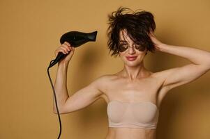 Attractive dark-haired woman with smoothing patches under eyes, holding hair dyer and drying her wet hair, making stylish hairstyle and cutely smiling looking at camera, isolated on beige background photo