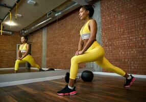 Side view of an African American beautiful athlete woman with perfect healthy slim body working out, stretching legs and performing lunges in gym club. Active lifestyle concept. photo