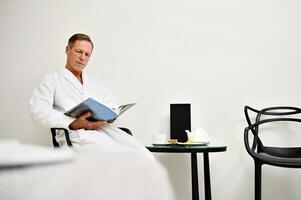 Handsome charming attractive mature Caucasian man in white terry bathrobe sitting on armchair and reading magazine, relaxing in private room at luxurious wellness spa centre photo