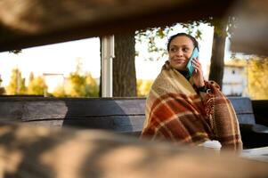 View through wooden logs of bench at beautiful woman looking to the side while talking on mobile phone, sitting on wooden bench wrapped in cozy checkered woolen blanket keeping warm on cool autumn day photo