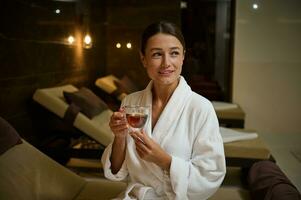Beautiful elegant Caucasian young woman in white bathrobe, holding a glass cup of hot healthy herbal drink, looking aside, sitting on chaise lounge by jacuzzi, enjoying spa procedures in spa salon photo