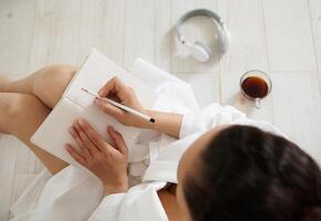 Top view of a brunette woman sitting on the floor and writing in a notebook. Cup with coffee and headphones lie on the floor photo