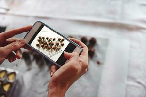 Closeup of hands holding mobile phone and making photography of delicious chocolate pralines on a white tablecloth. Mobile phone in live view regime. photo