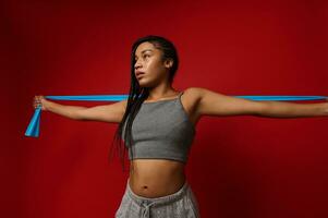 Determined young African woman with stylish dreadlocks stretching her arms using elastic rubber fitness band during routine workout against colored background with copy space photo