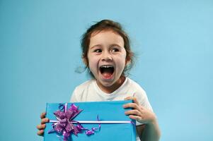 Cute little girl laughing while posing on blue background with gift in her hands. Copy space. Celebration concept. photo