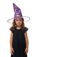 Portrait of pretty little girl wearing a wizard hat and dressed in stylish carnival dress, looking at camera posing with crossed arms against white background, copy space. Halloween concept photo