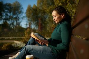 Attractive woman reading book while resting on wooden bench, enjoying warm sunny autumn weekend in country park, away from the hustle and bustle of the city, resting from digital gadgets and work photo