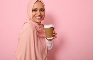 Close-up. Arabic Muslim woman with covered head in hijab holds disposable cardboard takeaway cup, smiles toothy smile, looking at camera, standing three quarters against colored background, copy space photo