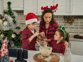 contento adorable niños en rojo y verde a cuadros ropa Cocinando con su mamá, haciendo magdalena masa, grabación vídeo para Navidad Cocinando vídeo Blog. alegre familia Cocinando juntos a hogar cocina foto