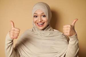 Happy Arab muslim woman in hijab with a beautiful toothy smile showing thumbs up to camera. Beige background with copy space. photo