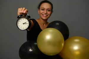 Focus on the hand holding a black alarm clock. Blurred smiling woman with gold black ir balloons gray background with copy space for advertisement Shopping at Black Friday photo