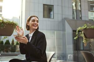 Attractive cheerful freelancer woman in business casual clothes sits on a summer terrace of a street cafe and smiles with a toothy smile, looking over her shoulder, holding a mobile phone in her hands photo