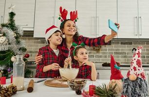 Cheerful family of beautiful mom in Santa hat and her adorable children, son and daughter talking by video call, making selfie, recording video, cooking Christmas bread together in the home kitchen photo