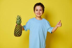 Handsome child boy holding a pineapple and showing thumb up while posing over yellow background with copy space photo