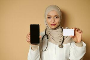 Confident portrait of a young Muslim female doctor holding a white plastic blank card and smartphone and showing them to the camera. Insurance concept photo