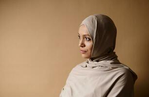 Closeup shot with soft shadow of serene elegant Muslim woman in traditional strict clothing looking the side on beige background with copy space photo