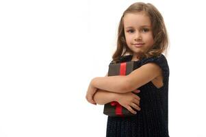 Beautiful 4 years old adorable baby girl gently hugs a black gift box and cutely looks at the camera, standing isolated against white background with copy space. Anniversary and Black Friday concept photo