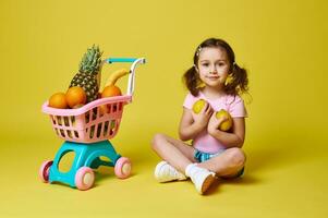 Adorable little girl sitting near a shopping cart full of fruits, holding two lemons in hands and cute smiling at camera . Isolated on yellow background with copy space photo