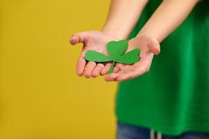 Closeup of hands holding a green clover leaf on the background of a boy standing on yellow surface with copy space. Saint Patrick concept photo