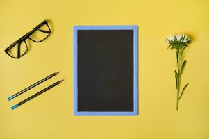 Top view. Flat lay. Autumn aster flower clean chalkboard with copy space , eyeglasses and two pencils, isolated on yellow background with space for text photo