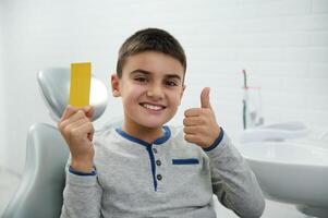 Confident handsome child boy holds yellow visit card with copy ad space, shows thumb up and smiles with beautiful toothy smile looking at camera while sitting in dentist's chair after dental treatment photo