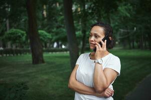 Confident multi-ethnic young pretty woman talks on mobile phone while strolling the alley of a city park on a summer day photo